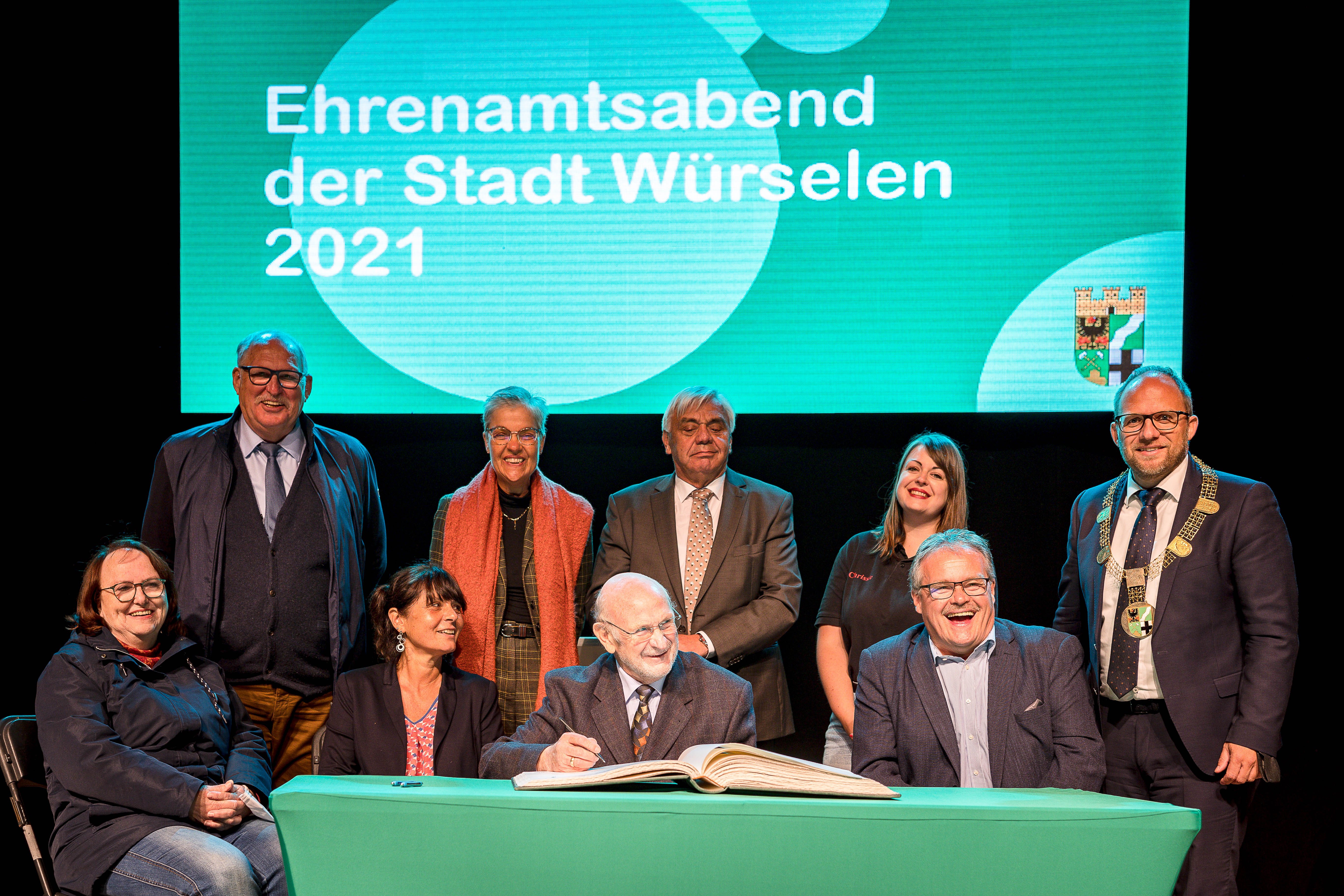 Neun Personen sitzen und stehen an einem Tisch und tragen sich in das Goldene Buch der Stadt Würselene ein