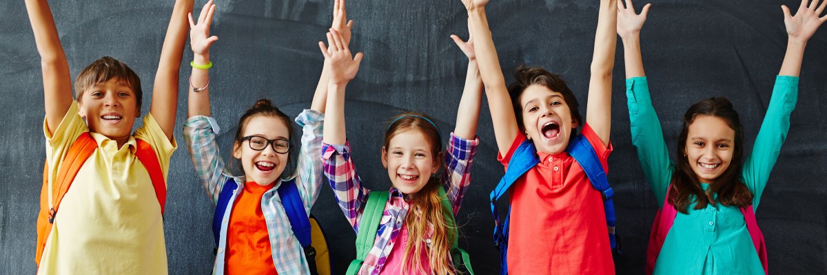 Glückliche Schulkinder stehen vor einer Tafel und strecken die Hände in die Höhe.