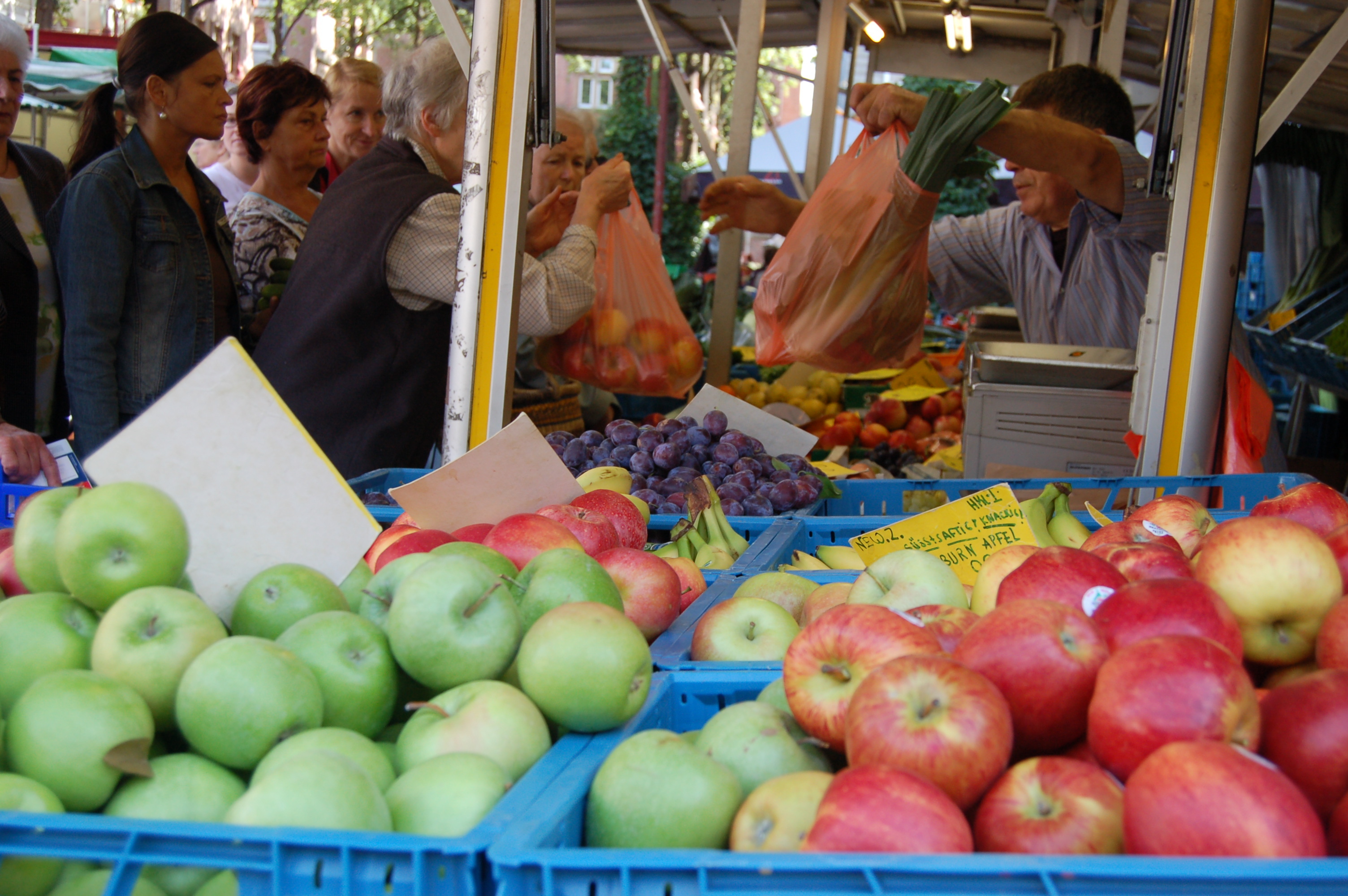 Kisten mit Obst an einem Marktstand, im Hintergrund Menschen, die einkaufen.