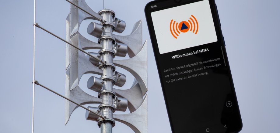 Nahaufnahme einer Sirene an einer Schule, daneben die geöffnete Warn-App NINA auf einem Smartphone