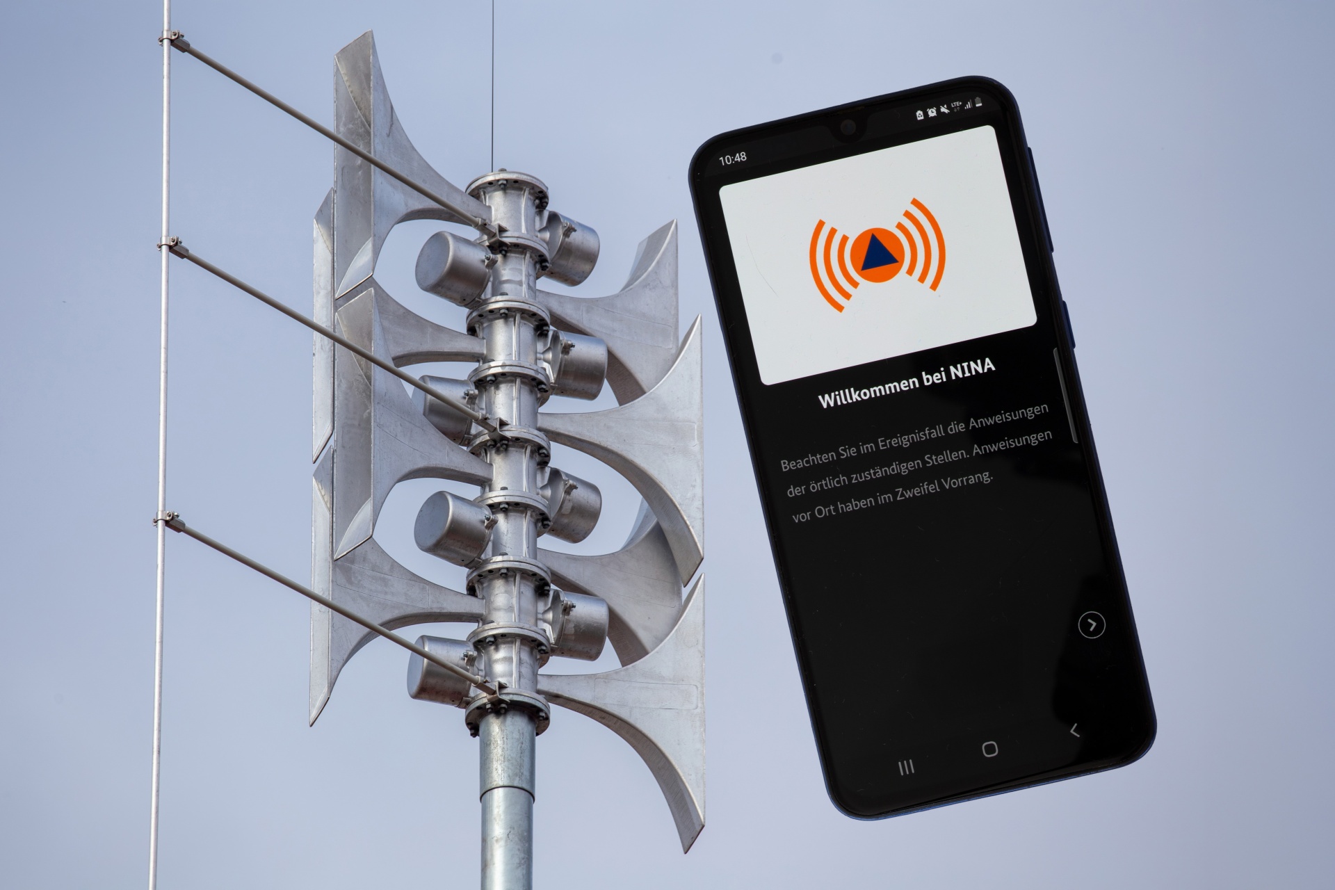 Nahaufnahme einer Sirene an einer Schule, daneben die geöffnete Warn-App NINA auf einem Smartphone