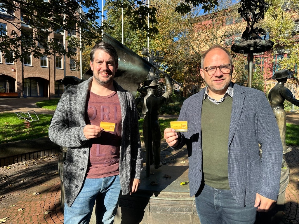 Bürgermeister Nießen und Mitarbeiter Volker Deutmann halten die Ehrenamtskarte in die Kamera.