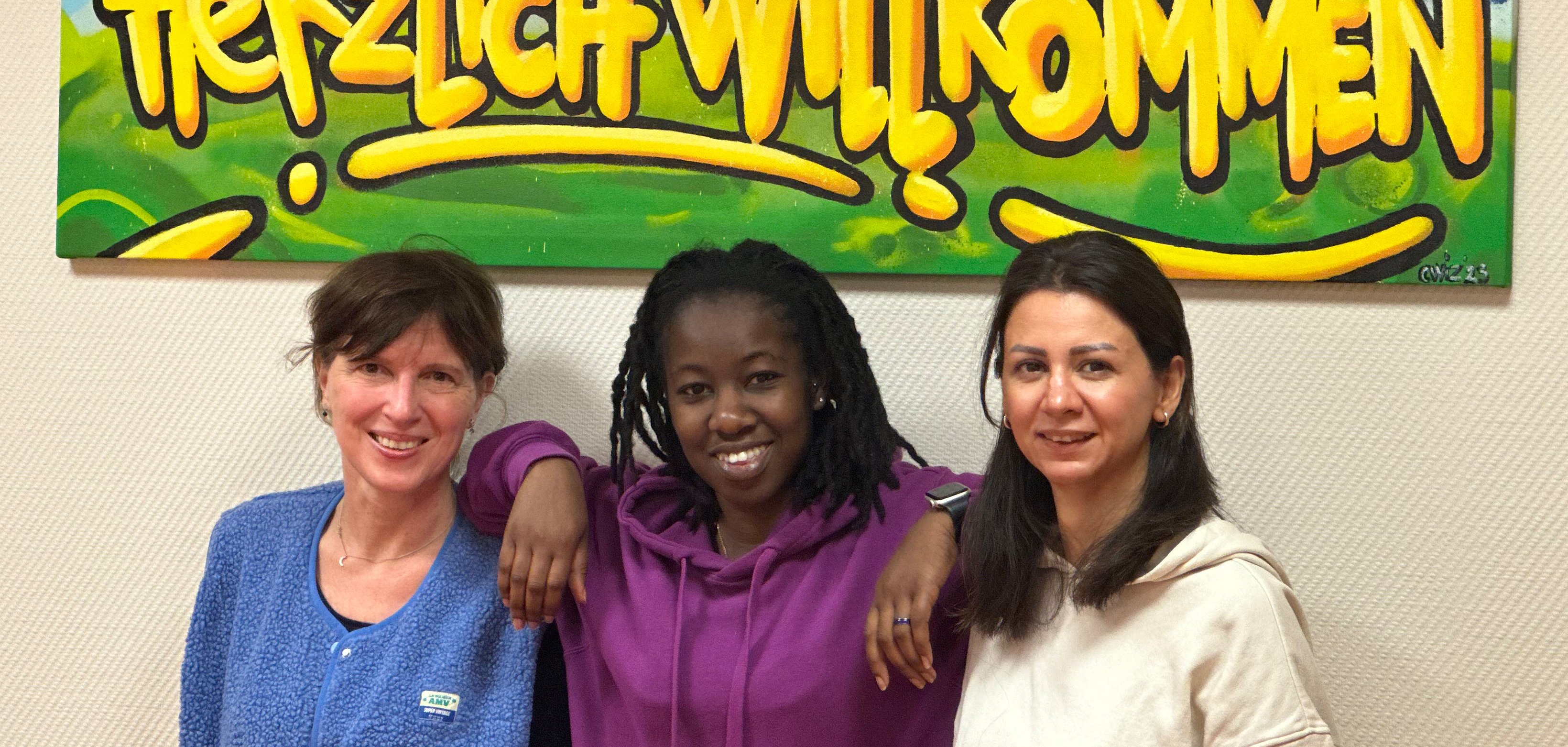 Drei Frauen stehen vor einer Wand. Im Hintergrund ein Schild mit Schriftzug "Herzlich Willkommen"
