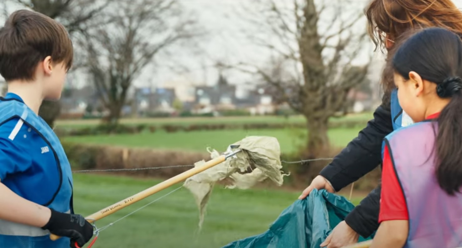 Ein Junge führt mit einer Greifzange Müll in einen Müllsack, eine Frau hält den Sack fest, ein Mädchen schaut zu,