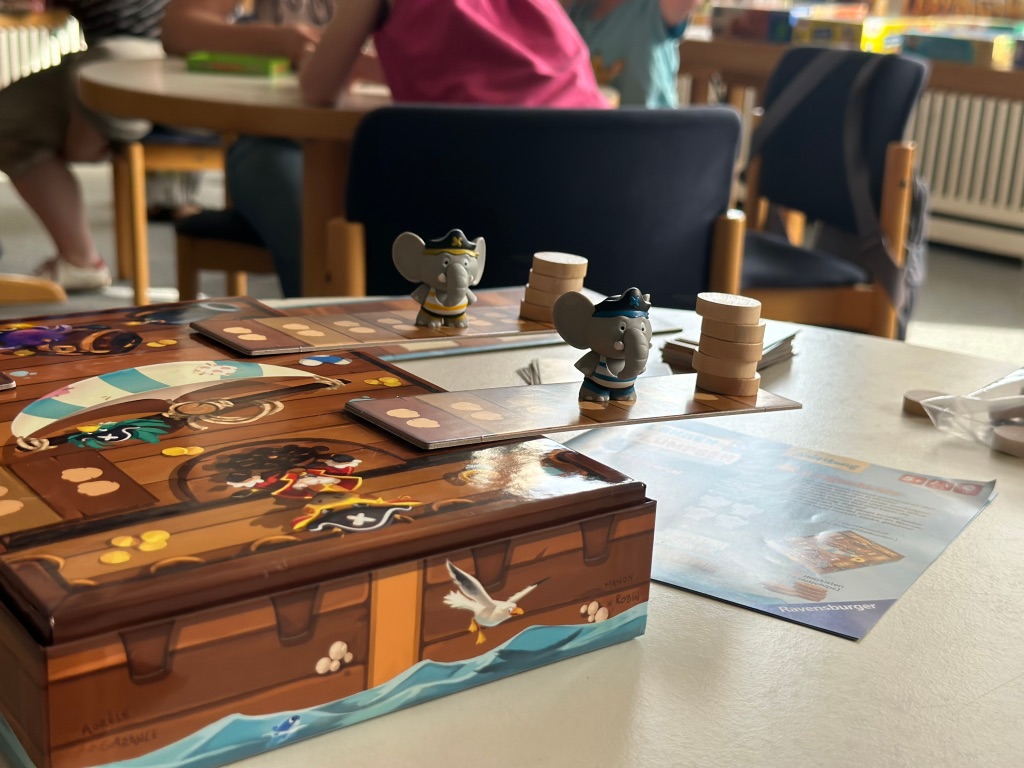 Brettspiel im Vordergrund, dahinter spielende Familien.