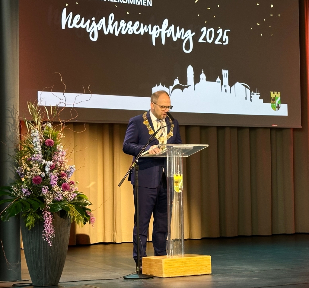 Der Bürgermeister steht auf der Bühne an einem Rednerpult und spricht zu den Leuten im Publikum.