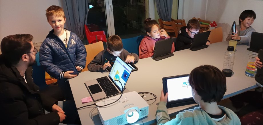 Mehrere Kinder sitzen an einem Tisch und arbeiten mit Computer, Tablet und Beamer