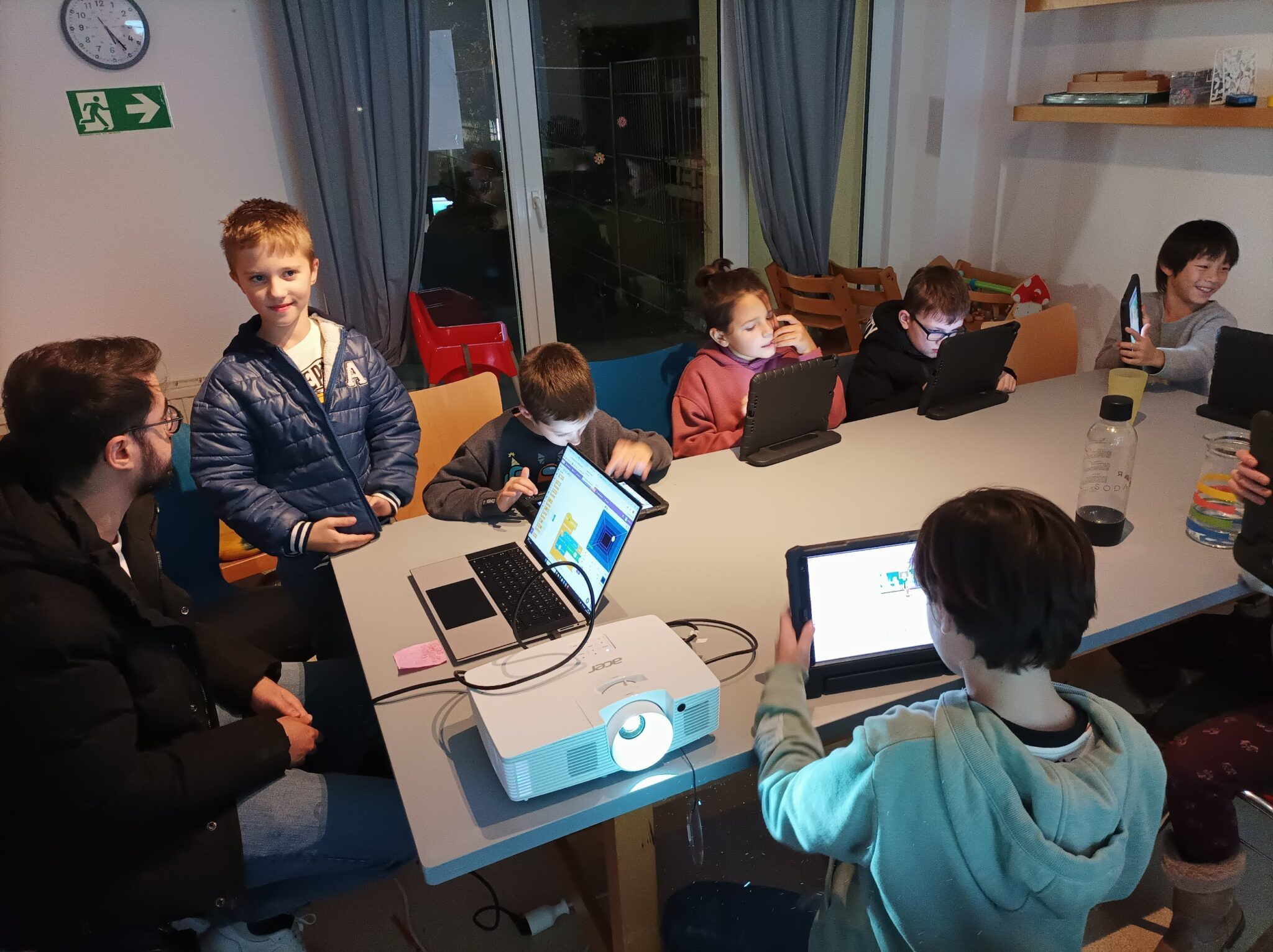 Mehrere Kinder sitzen an einem Tisch und arbeiten mit Computer, Tablet und Beamer