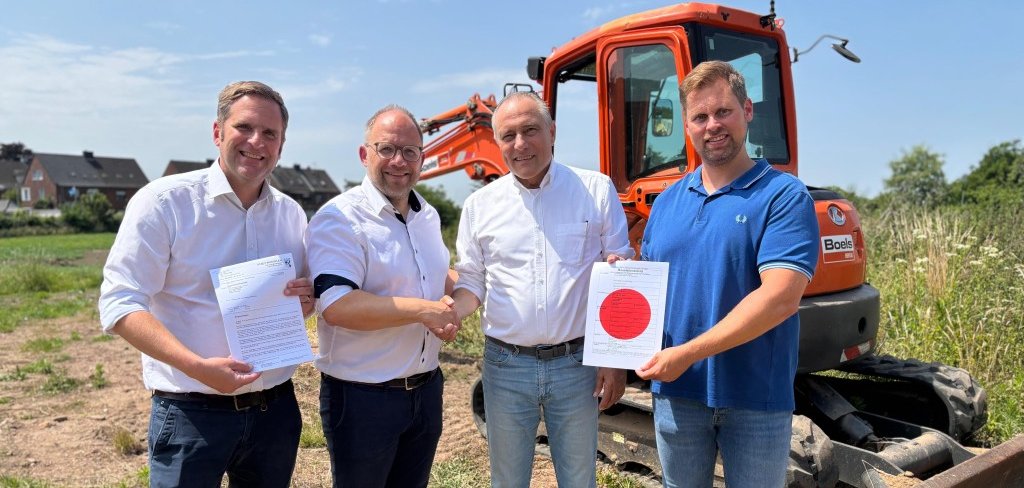 Vier Herren vor einem Bagger, in den Händen einen Schriftstück und ein Baustellenschild.