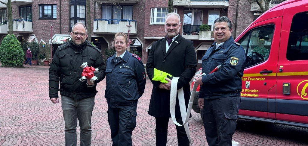 Vier Leute halten Feuerwehr-Gegenstände in den Händen, vor ihnen liegen weitere Sachen auf dem Boden, hinter ihnen steht ein Feuerwehrauto.