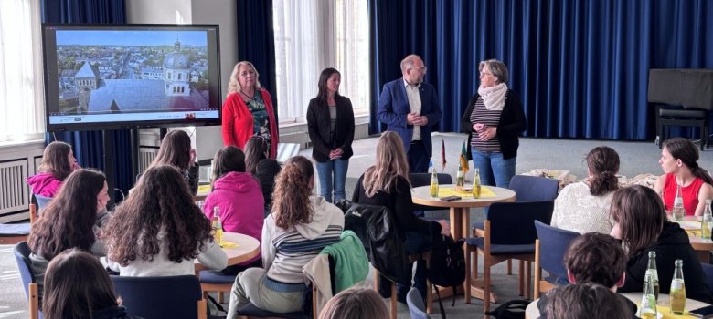 Französische Schüler im Alten Rathaus (Lehrer) Vier Erwachsene sprechen, davor sitzen Schüler an Tischen und hören zu.