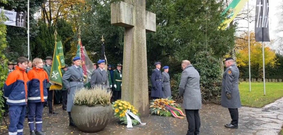 Einige Menschen mit Flaggen am großen, steinernen Kreuz auf dem Friedhof, davor zwei Trauerkränze