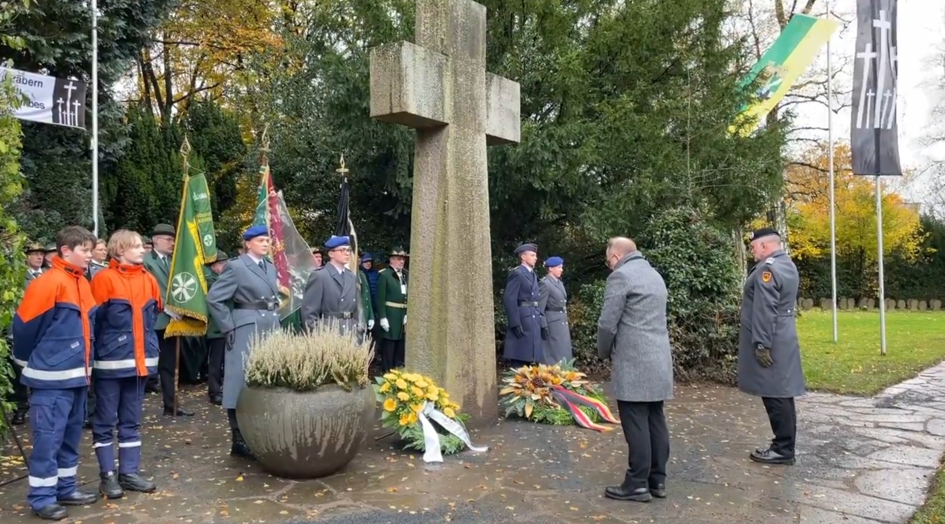 Einige Menschen mit Flaggen am großen, steinernen Kreuz auf dem Friedhof, davor zwei Trauerkränze