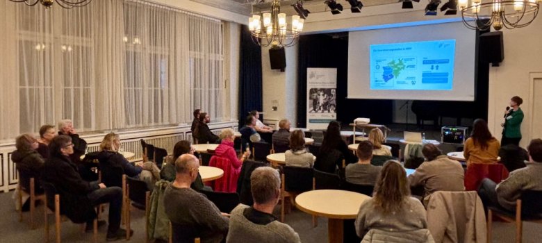 Ein Saal mit Menschen, die zu einer Bühne schauen, wo eine Präsentation gezeigt wird.