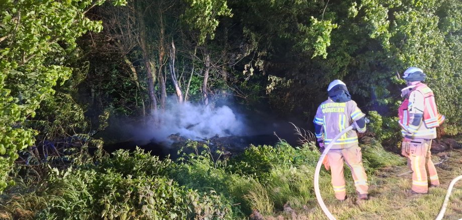 Zwei Einsatzkräfte der Feuerwehr Würselen löschen einen Brand in einer Böschung, aus der weißer Rauch aufsteigt. Die Einsatzstelle im Gebüsch wird dabei von Scheinwerfern hell ausgeleuchtet.