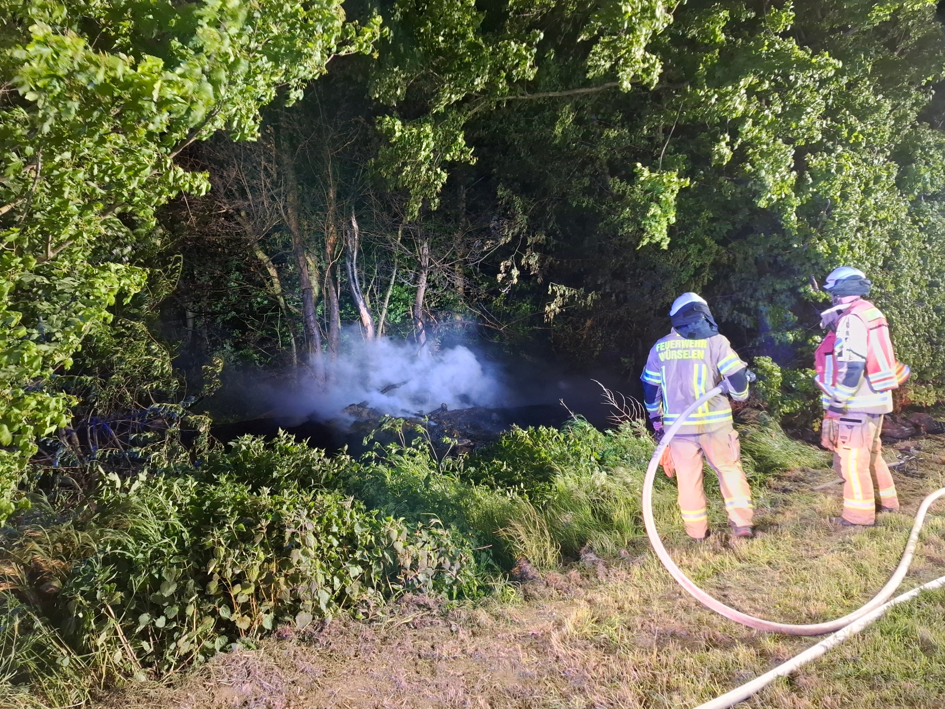 Zwei Einsatzkräfte der Feuerwehr Würselen löschen einen Brand in einer Böschung, aus der weißer Rauch aufsteigt. Die Einsatzstelle im Gebüsch wird dabei von Scheinwerfern hell ausgeleuchtet.
