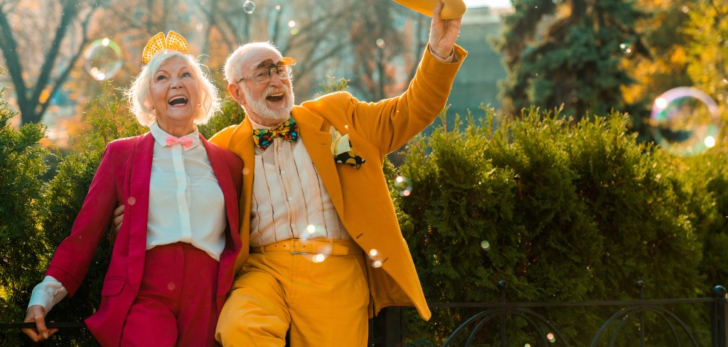 Funny elderly woman laughing in the park next to her husband Ein glückliches älteres Pärchen in bunten Kostümen tanzt fröhlich im freien.