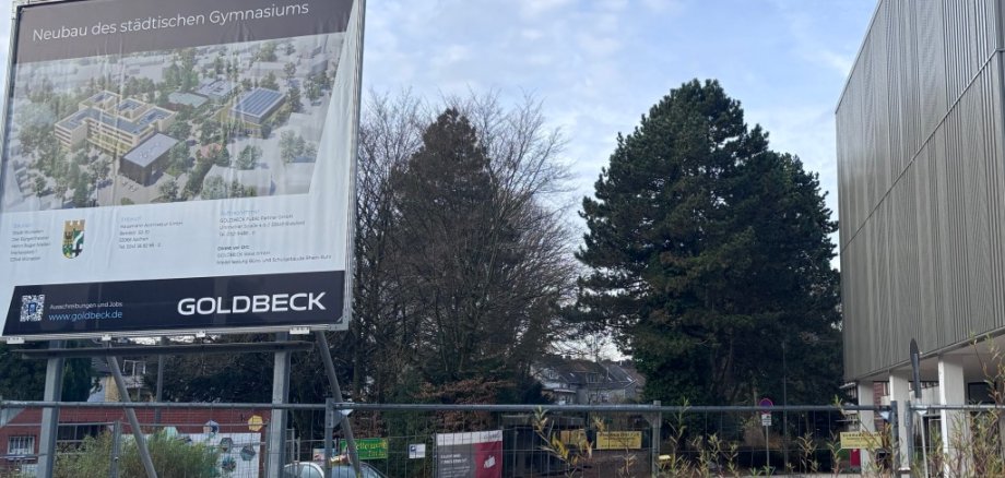 Baustellenschild mit Informationen zum Neubau des Gymnasiums, im Hintergrund die Aula/Stadthalle und Teile des alten Gebäudekomplexes