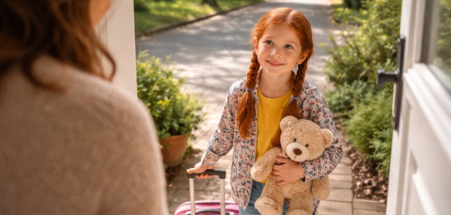 Ein kleines Mädchen mit einem Teddybären und einem Koffer steht lächelnd vor einer geöffneten Haustüre. Eine Frau erwartet Sie dort bereits.
