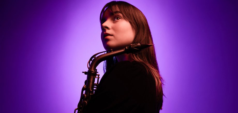 Paula Steimer Paula Steimer. Eine junge Frau mit dunklem Haar und einem Saxofon steht seitlich vor einem farbigen Hintergrund